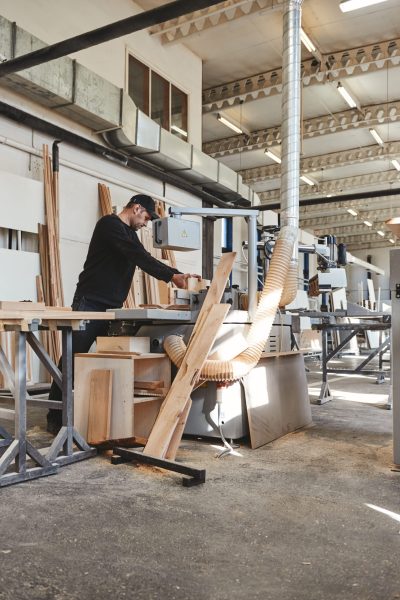 Skilled carpenter in black uniform cutting a piece of wood in his woodwork workshop. Other machinery in the background. Vertical shot